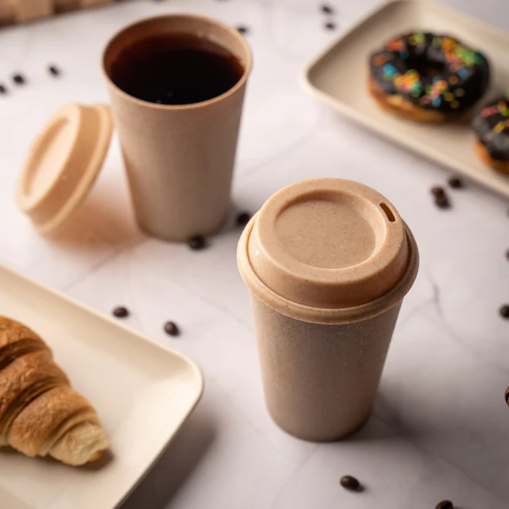 Eco-friendly travel mug with a coffee cup, lid on, croissant, and donuts on a marbled surface, with coffee beans around.