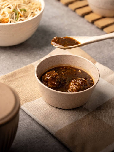 A rice husk bowl containing curry with meatballs, with a spoonful being lifted, set on a textured tablecloth alongside another bowl of noodles.