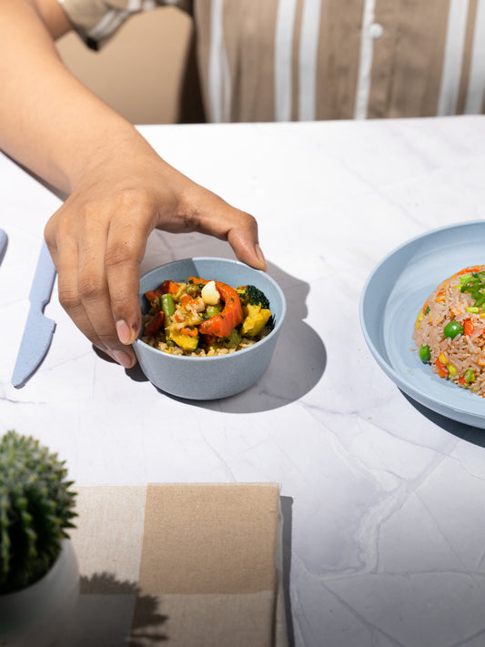 Colourful vegetable curry in a wheat straw ramen bowl with an Irida wooden spoon on a teal napkin, surrounded by assorted dishes in rice husk and wheat straw dinnerware.