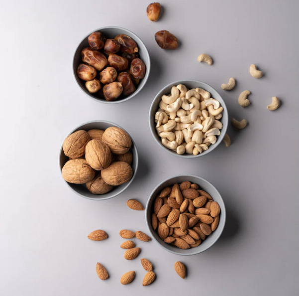 Four bowls with walnuts, almonds, and cashews, alongside scattered nuts and dates on a grey background.