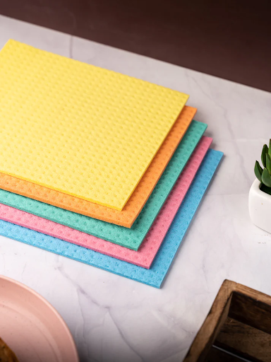Stacked colorful sponge wipes on a marble surface next to a plant and wooden tray, showcasing hygiene and organization.