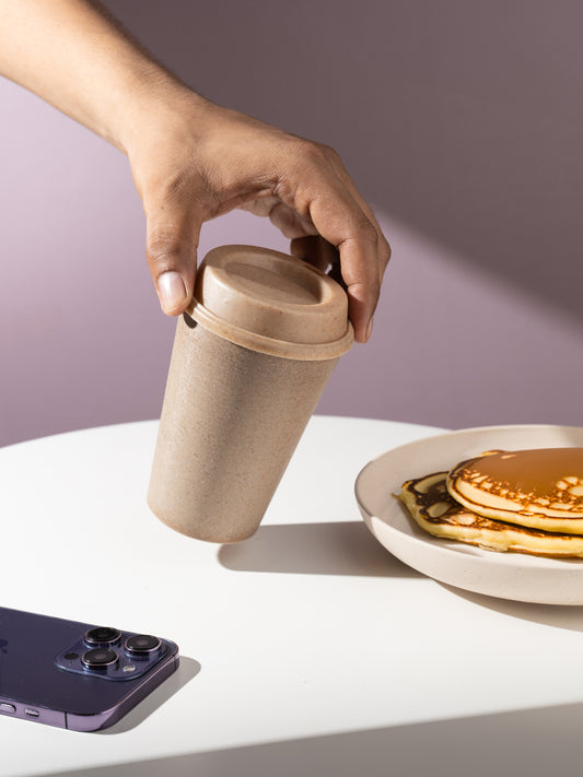 A hand reaching for an eco-friendly, reusable coffee cup with a sipper lid on a white table, next to a plate of pancakes with syrup and a purple smartphone.