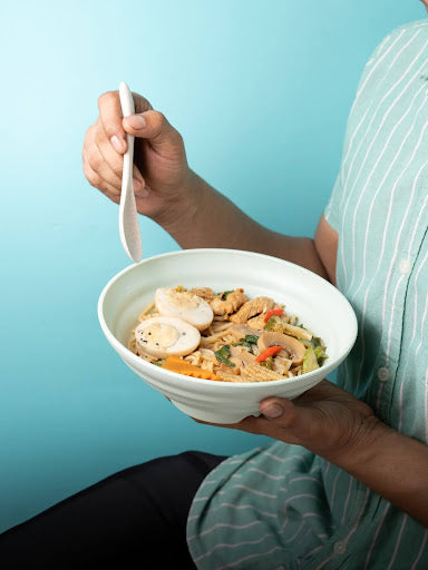A person in a striped green and white shirt holding a wheat straw bowl filled with stir-fried noodles, vegetables, and sliced boiled eggs. The bowl is held up against a turquoise background.