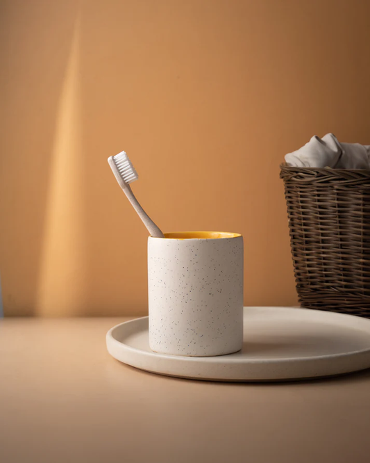 A disposable toothbrush in a ceramic holder on a tray, with a basket of linens in the background, simple and elegant.