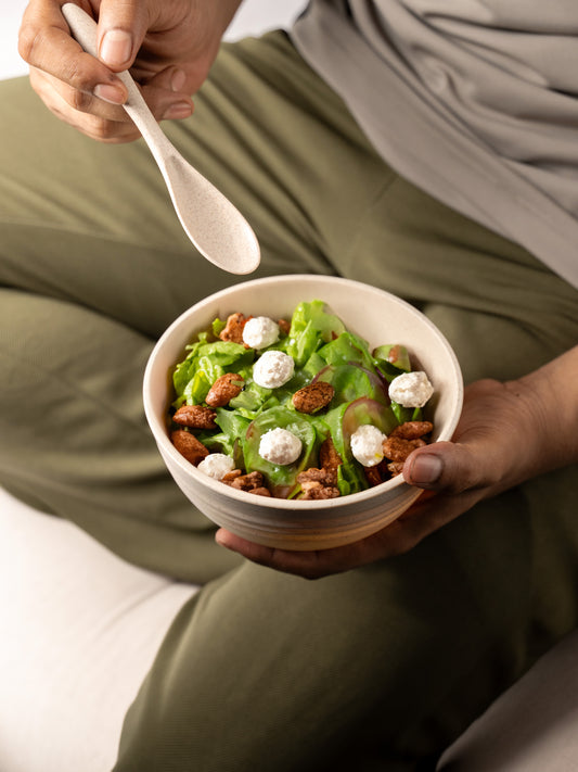 Person seated in olive green pants and a gray top, holding a wheat straw bowl filled with a vibrant green salad with cheese and walnuts, using a wheat straw spoon, set against a soft white background.