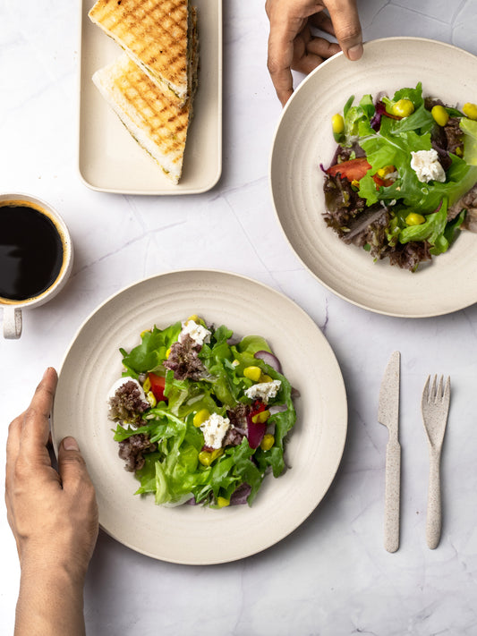 A top-down view of a table setting with two plates of green salad with cheese, a cup of black coffee, toasted sandwiches on a rectangular plate, and a person's hand reaching for a fork.