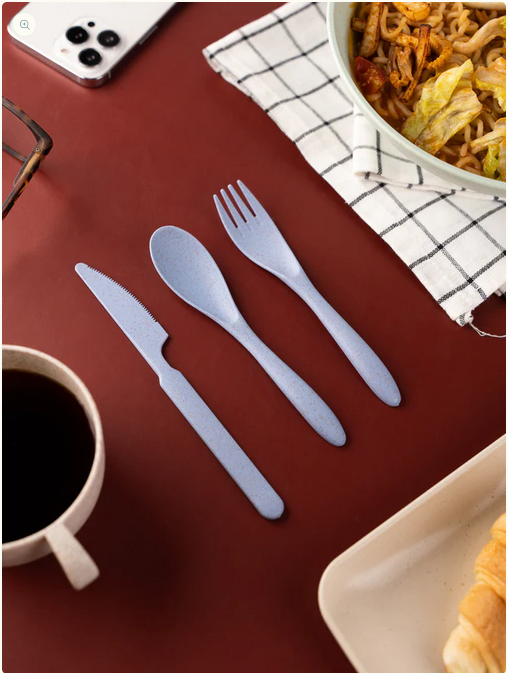 A casual dining setup with eco-friendly cutlery on a burgundy table, alongside a cup of coffee, a bowl of noodles, a croissant, and a smartphone with a checkered napkin.