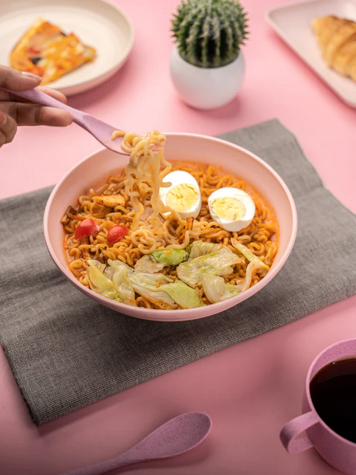 A person's hand using a pink fork to lift noodles from a pink bowl full of ramen with boiled eggs and vegetables, on a pink table with a grey napkin, a coffee cup, and other dishes in the background.