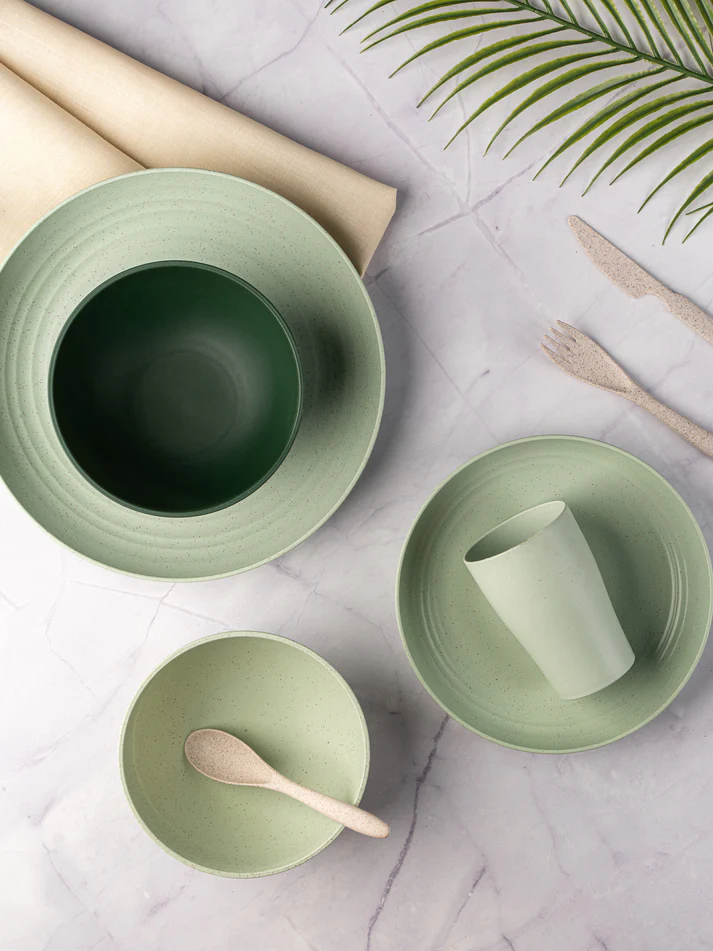 SustainableWheat Straw tableware in mint green, with a cup, bowls, and cutlery on a marble background, beside a palm leaf.
