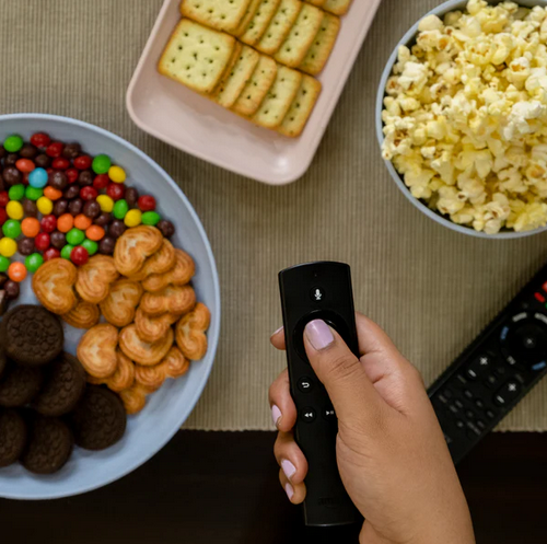 A person's hand holding a TV remote, with eco-friendly plates of snacks including crackers, candies, cookies, and popcorn, and another remote control on a textured brown surface.