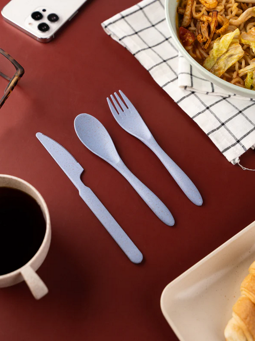 Eco-friendly cutlery laid out on a burgundy table, with a cup of coffee, a bowl of noodles, a croissant, a checkered napkin, and a smartphone in the background.
