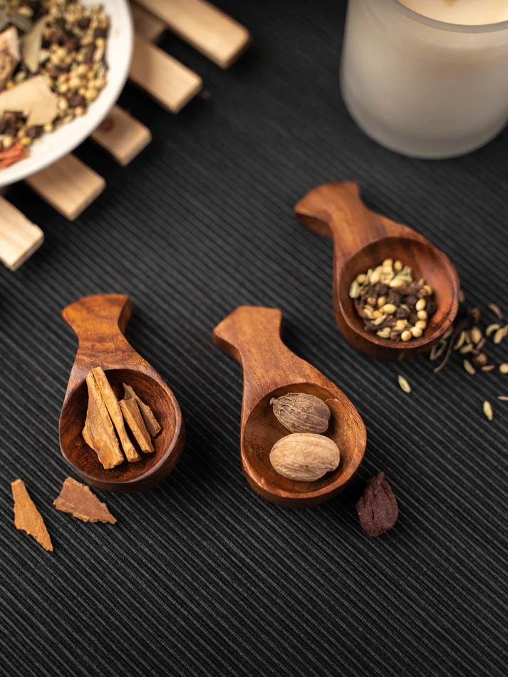 Wooden cutlery spoons with spices on a dark mat, near scattered herbs and a candle, invoking a cozy ambiance.