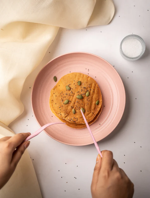 Hands cutting into pancakes on an eco-friendly pink plate, with a small bowl of sugar and a beige cloth napkin on a white table.