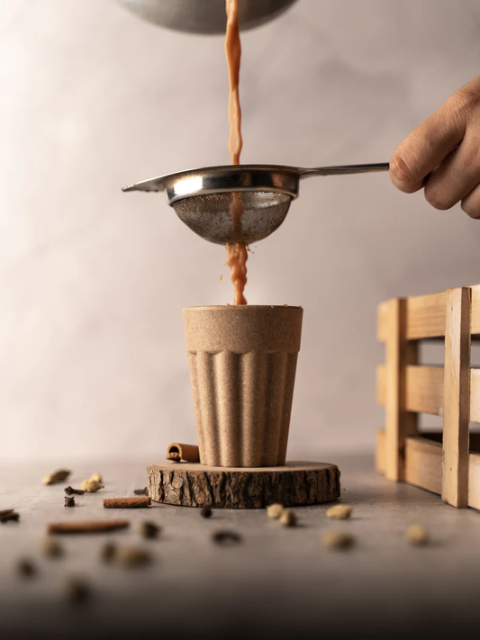 Pouring chai through a strainer into a rice husk cutting chai cup on a rustic wood slice, with spices scattered around.