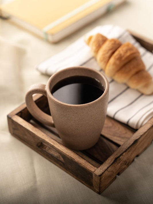 A wheat straw coffee mug filled with black coffee on a rustic wooden serving tray, accompanied by a freshly baked croissant on a striped napkin, with a yellow notebook blurred in the background, all arranged on a beige surface.