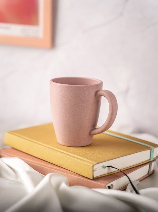 A pink eco-friendly mug resting on top of a yellow hardcover book, with a white fabric and a marble background, creating a cozy reading atmosphere.